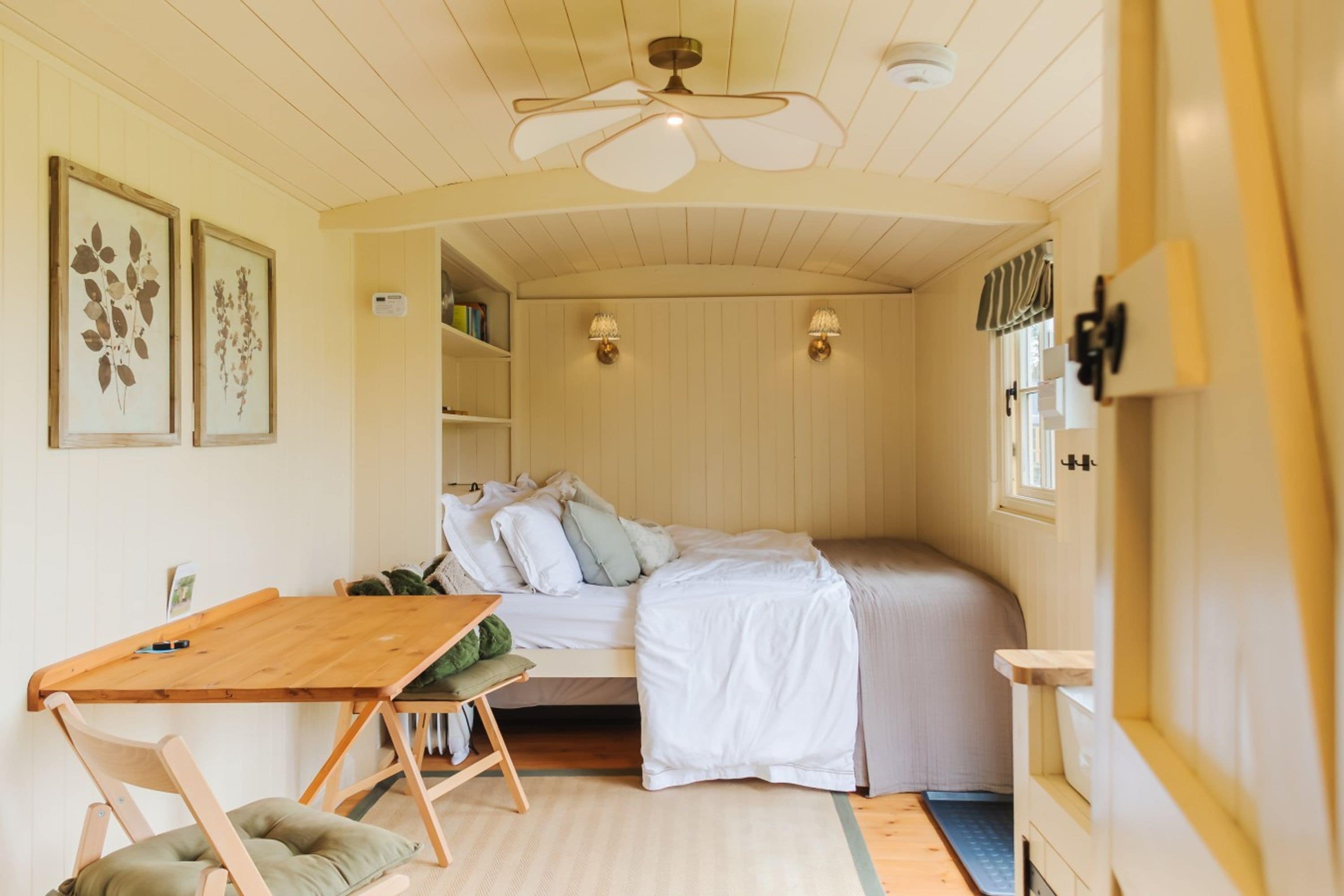 A compact room with a bed, a small table, and a chair. There are wall-mounted lights and a ceiling fan.