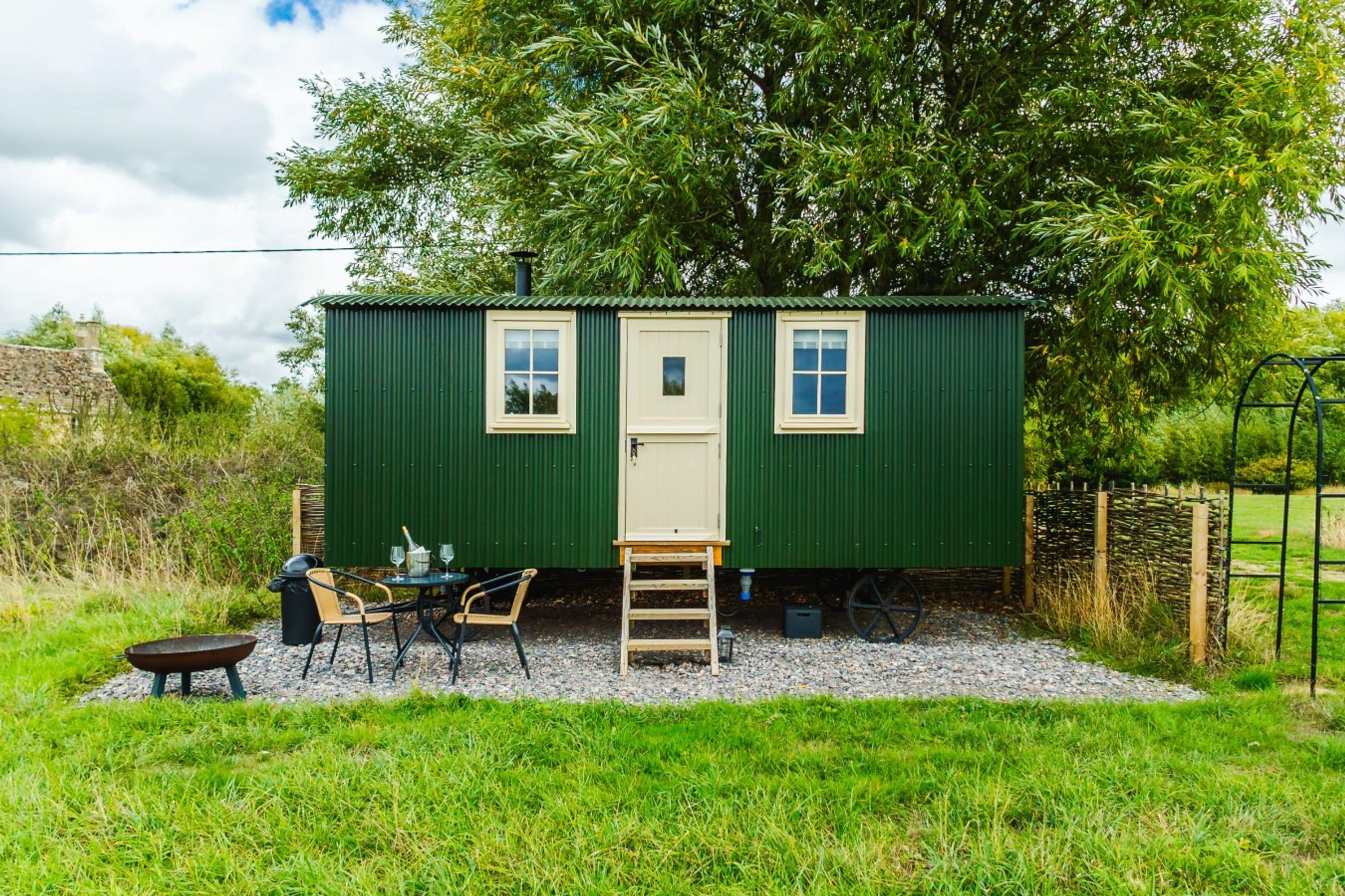A small green building with a door and two windows, a table with chairs, and a ladder outside.