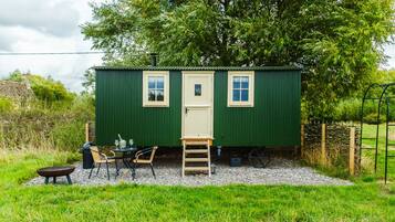 A small green building with a door and two windows, a table with chairs, and a ladder outside.