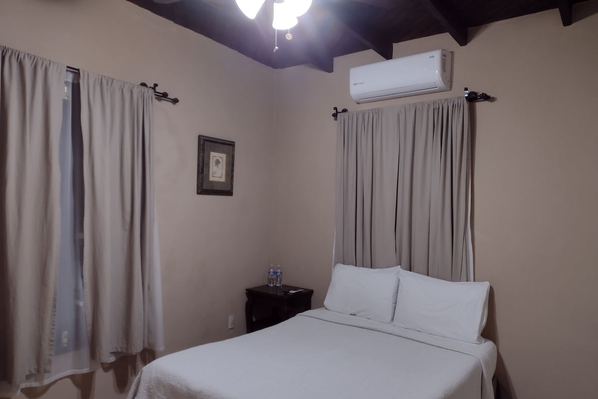 A bedroom with a bed, curtains, an air conditioning unit, a nightstand, and a framed picture on the wall.