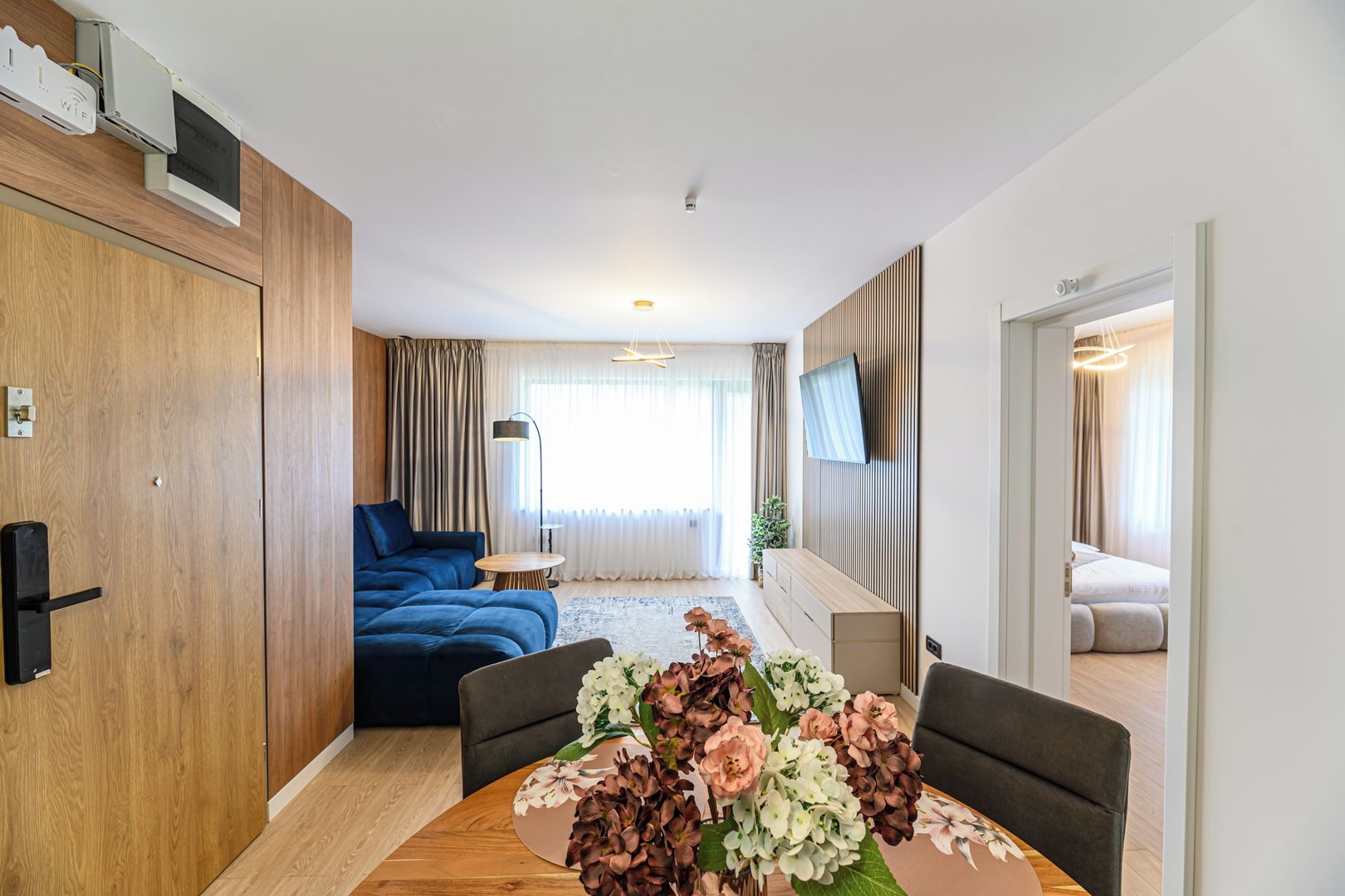 A modern living room with a sofa, a dining table with flowers, a TV mounted on the wall, and a wooden door.