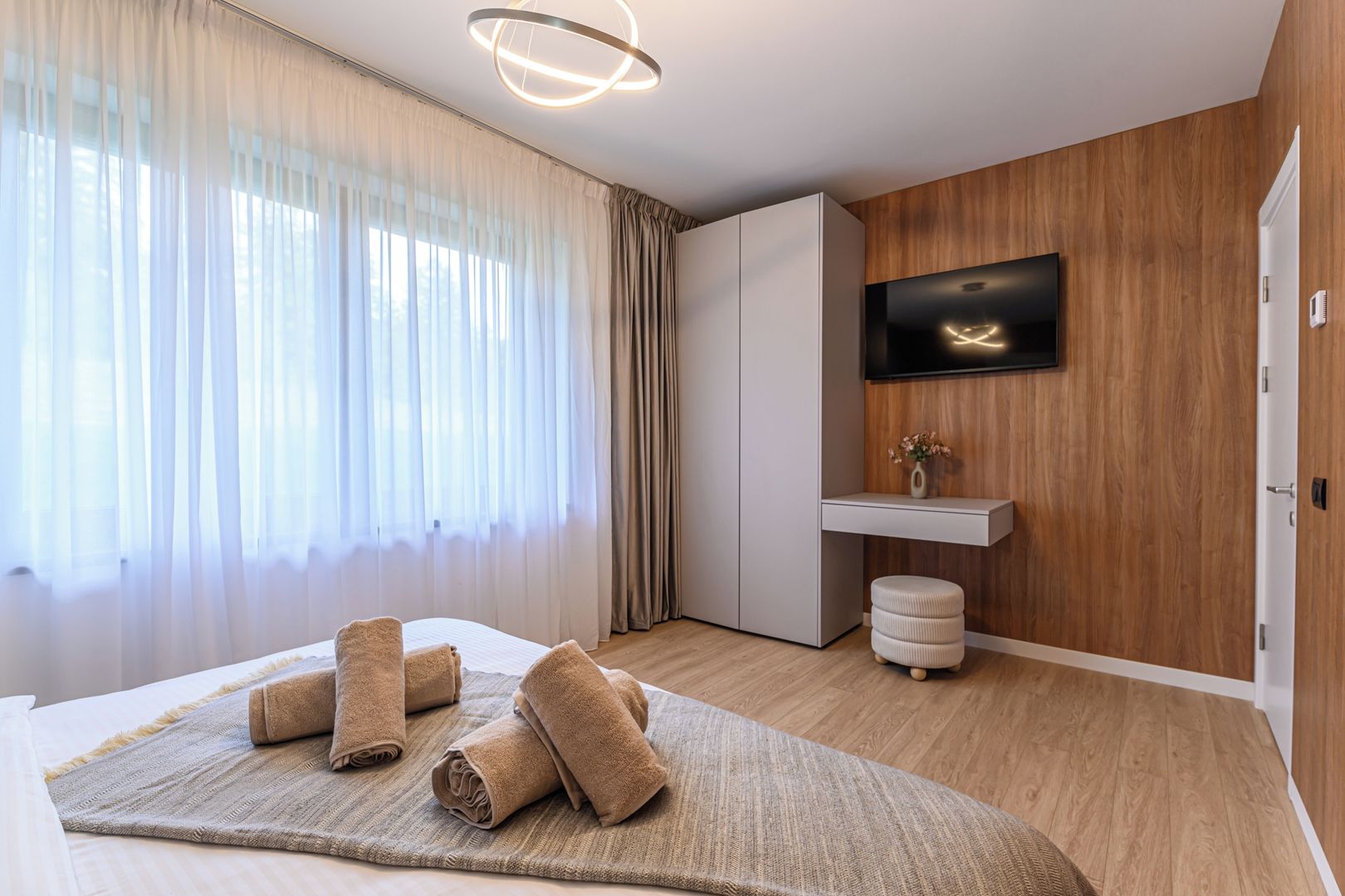 A modern hotel room with a bed, a bedside table, a flat-screen TV mounted on a wooden wall, and a large window with sheer curtains.