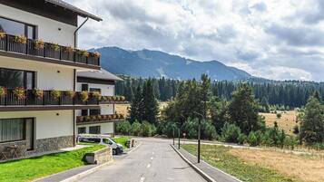 A multi-story building with balconies, a car parked in front, and a mountainous landscape in the background.