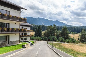 Un immeuble à plusieurs étages doté de balcons, une voiture garée devant, et un paysage montagneux en arrière-plan.