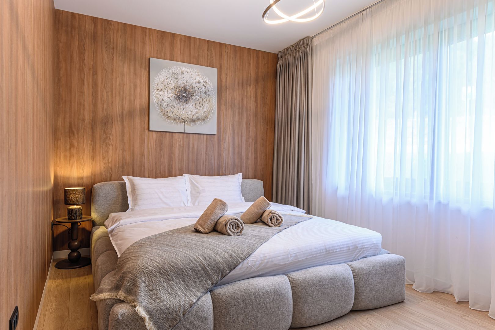 A modern bedroom with a large bed, a bedside lamp, a wall-mounted artwork, and sheer curtains.