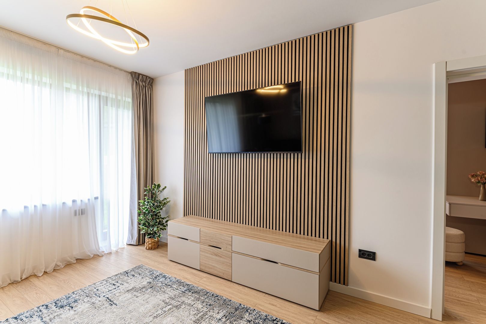 Ein modernes Wohnzimmer mit einem Flachbildfernseher an einer Wand aus Holzpaneelen, einer Holzkassette mit Schubladen, einer Topfpflanze und einem großen Teppich.
