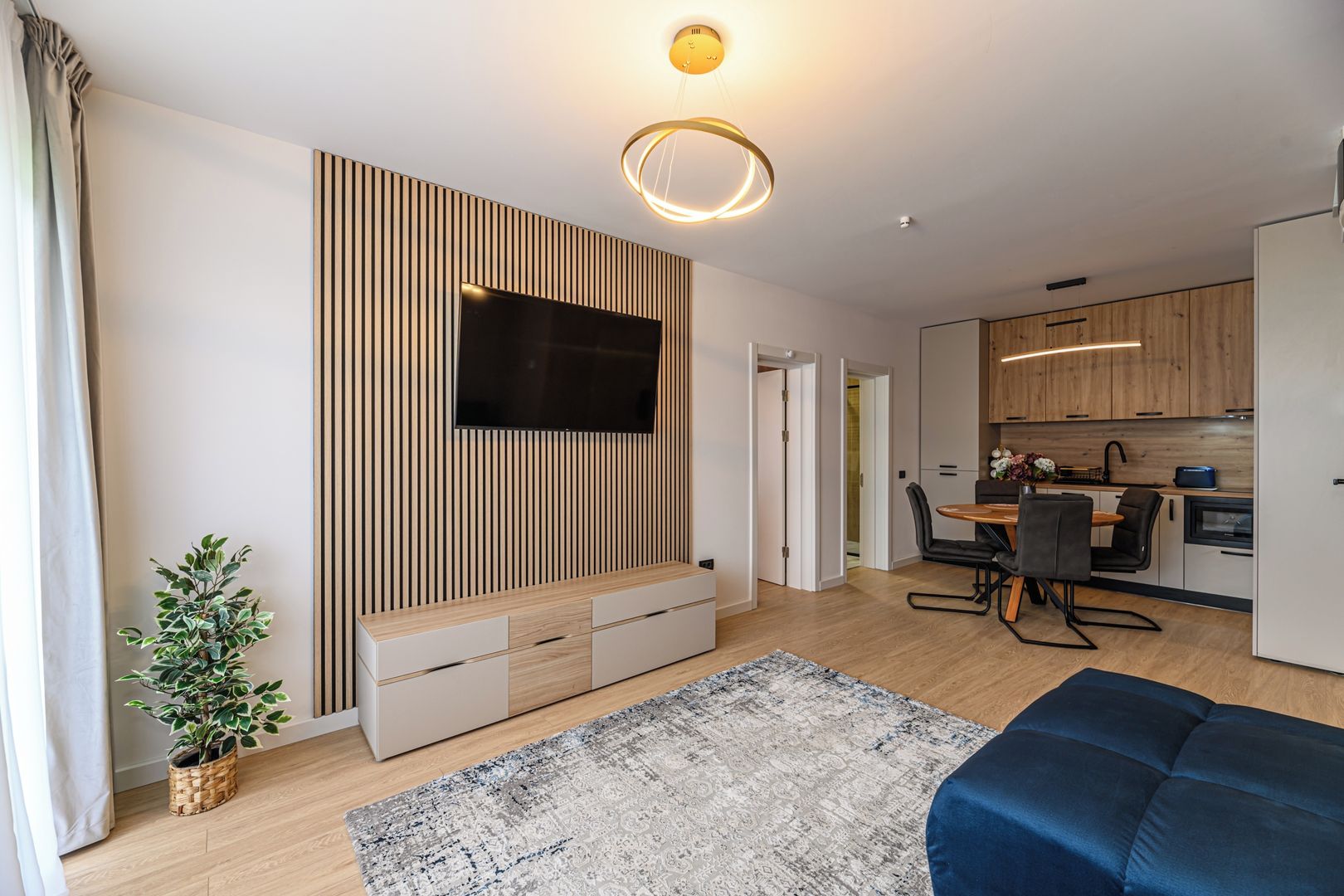 Uma sala de estar moderna com televisão de tela plana, uma parede decorativa de madeira, uma área de refeições com mesa e cadeiras, e uma cozinha ao fundo.