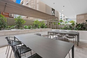 Ein Essbereich im Freien mit einem langen Tisch und Stühlen, umgeben von Grünpflanzen und einer Holz-Pergola.