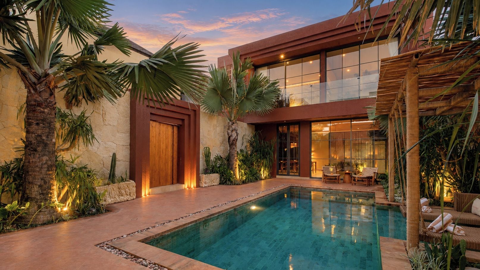 A modern pool area with a clear pool, surrounded by palm trees and plants, and a building with large windows and a wooden door.
