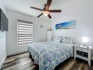 Ein Schlafzimmer mit einem Bett, einem Nachttisch, einem Deckenventilator, einem Fenster mit Jalousien, einem an der Wand montierten Fernseher und einem Bild einer Strandlandschaft.