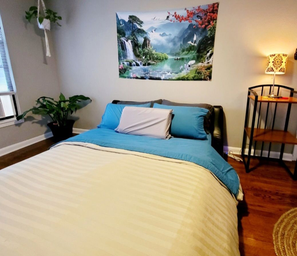 Un dormitorio con cama, un cuadro de un paisaje, una planta en maceta y una lámpara.
