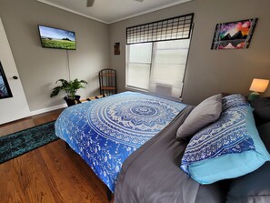 Een slaapkamer met een bed, een televisie, een raam met zonwering, een plant en een schilderij aan de muur.