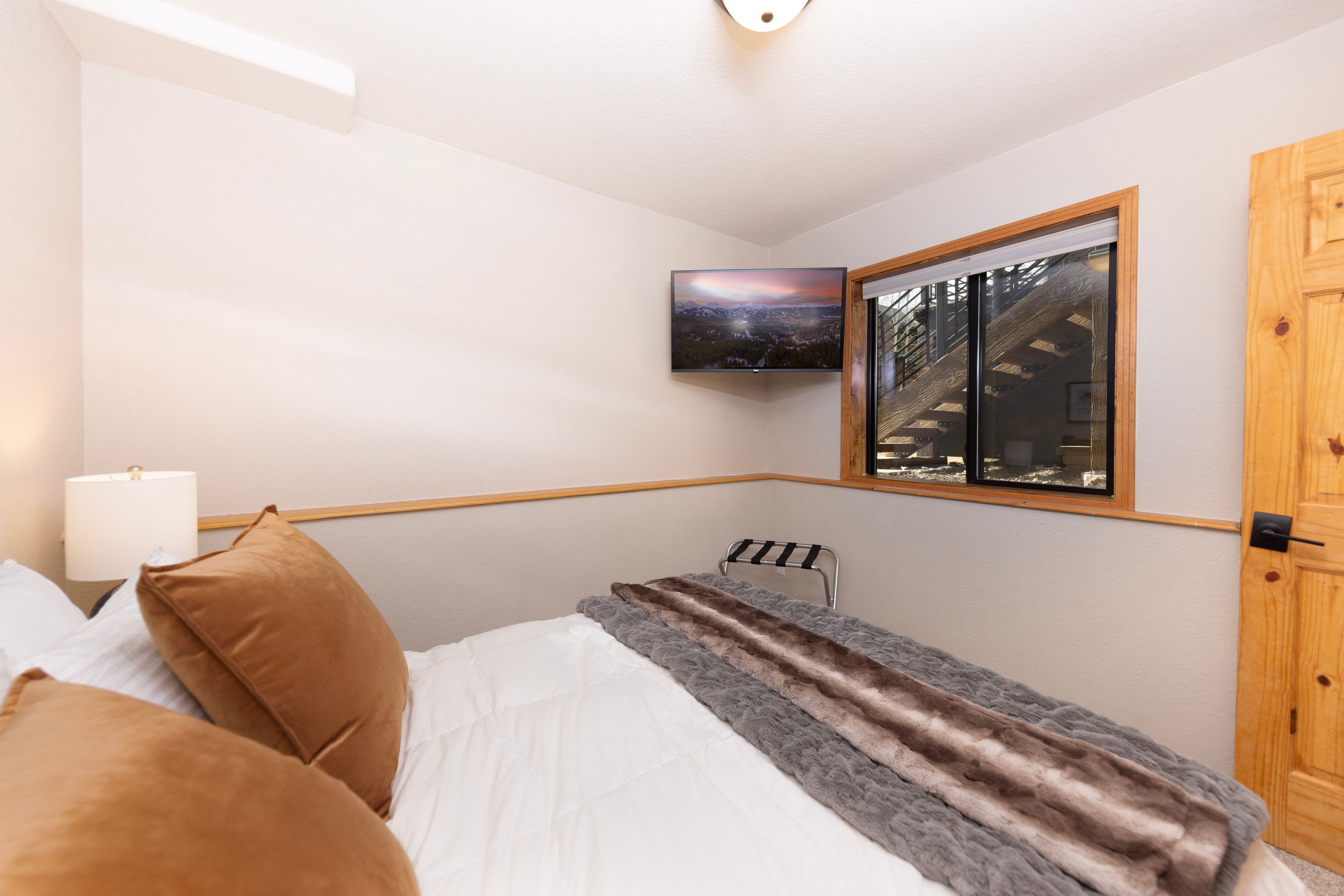 A bedroom with a bed, pillows, a TV mounted on the wall, and a window with a view of a building.