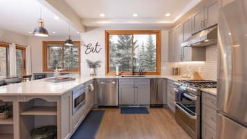 A modern kitchen with a central island, stainless steel appliances, and a view of trees through the windows.