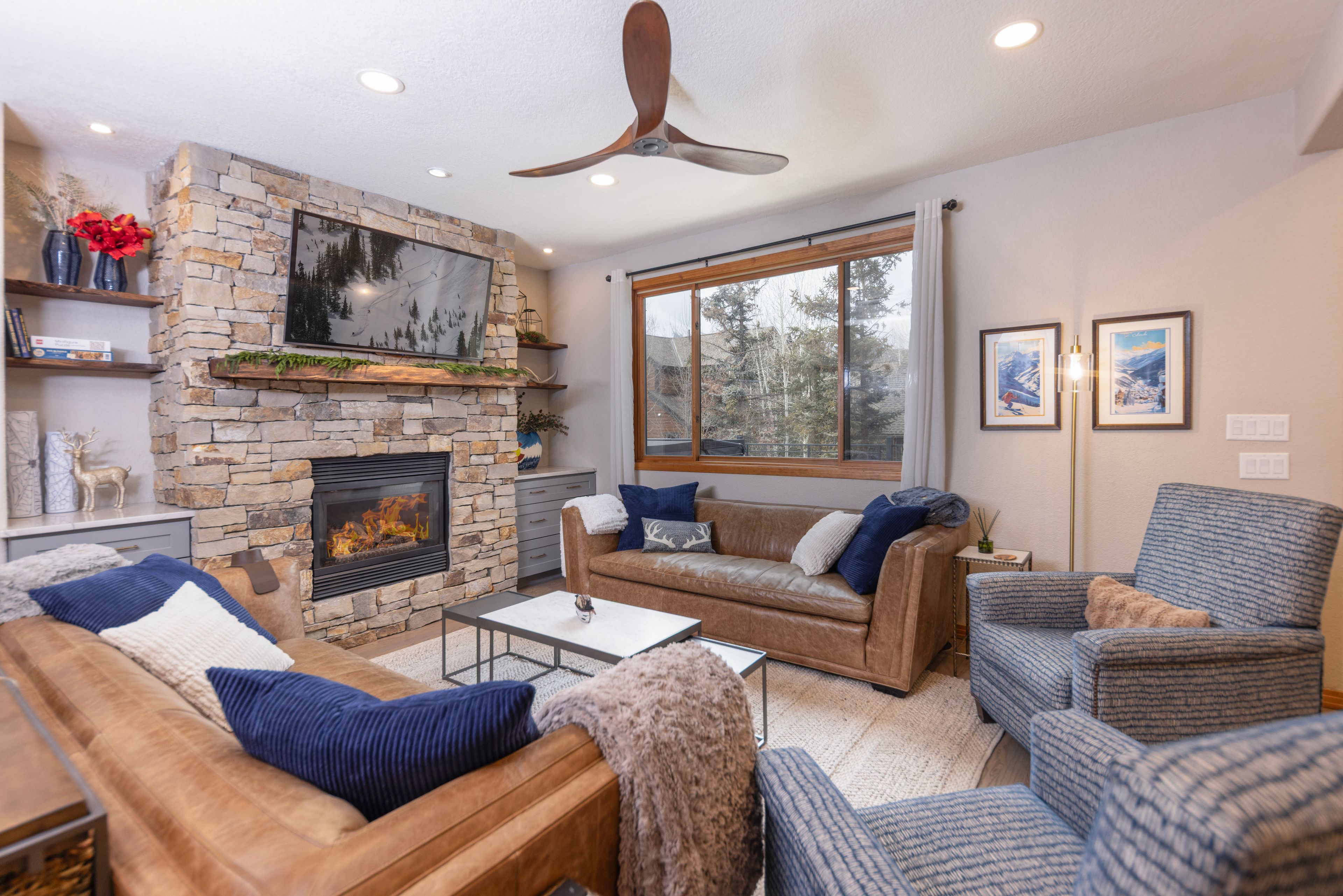A living room with a stone fireplace, leather sofas, a coffee table, and a large window with a view of trees.
