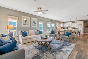 A spacious living room with a large sofa, a coffee table, and a dining area with chairs. There is a kitchen in the background with white cabinets and a ceiling fan. - Ocean View | Inflatable Hot Tub | Arcade Game (Crystal Beach)