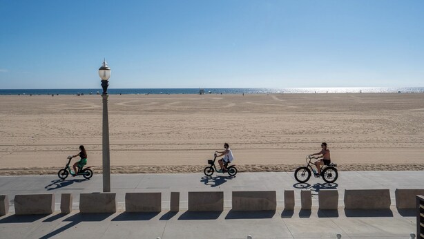 Una spiaggia con una passeggiata lastricata, un lampione e tre persone che vanno in bicicletta.