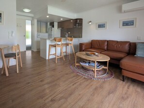 A modern living room with a brown leather sofa, a round coffee table with a woven base, and a dining area with wooden chairs and a small table. - Sunseeker Studio - Ground Floor Apt (Noosa Heads)