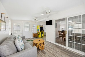 A living room with a sofa, a small round table, a ceiling fan, and a Pac-Man arcade game. - 3BR, 2BA home in Fernandina Beach's North End, on Amelia Island (Fernandina Beach)