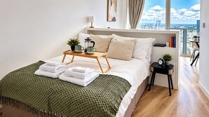 Una cama bien hecha con una manta verde y toallas blancas, una mesita de noche con una planta, una cafetera y libros.