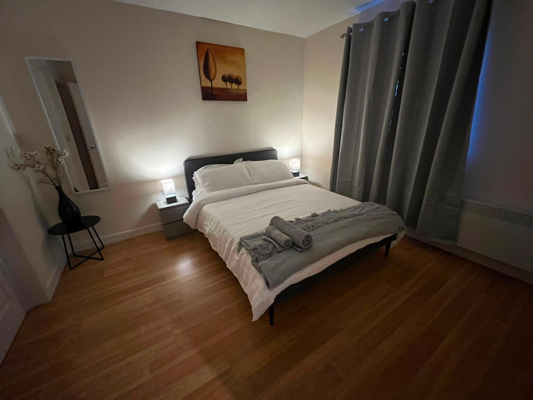 A hotel room with a bed, bedside tables, a nightstand, a vase with dried flowers, a painting on the wall, and a window with curtains.