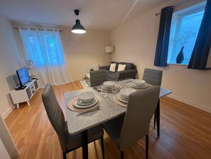 Un comedor con una mesa dispuesta para cuatro, un sofá, un televisor y una ventana con cortinas.