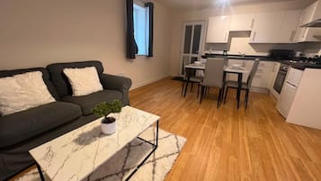 A modern living room with a sofa, a coffee table with a plant, and a dining area with chairs and a kitchen in the background.