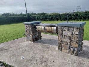 Eine Steinsitzbank mit einem Metallfass und einem Wasserhahn, auf einer Betonfläche mit Gras und einer Hecke im Hintergrund.