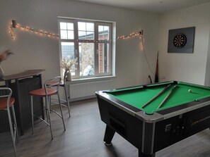 Ein Billardtisch, eine Dartscheibe, ein Barbereich mit Hockern und ein Fenster mit Blick nach draußen.