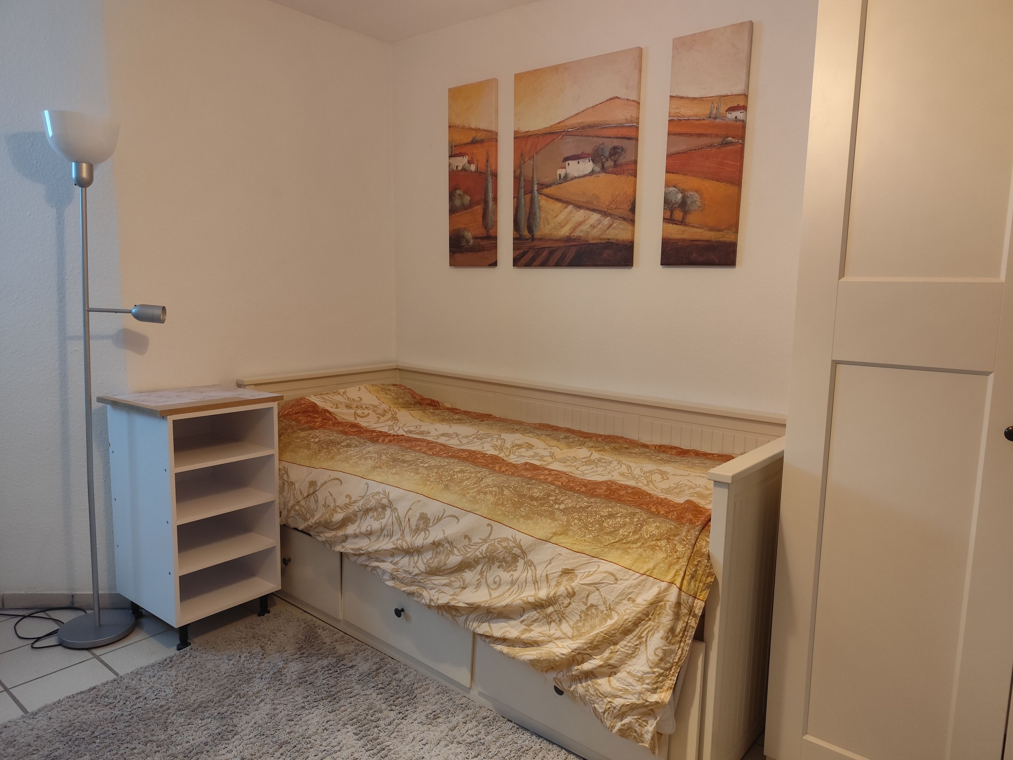 Ein kleines Schlafzimmer mit einem Einzelbett, einem Nachttisch, einer Lampe und drei Bildern an der Wand.