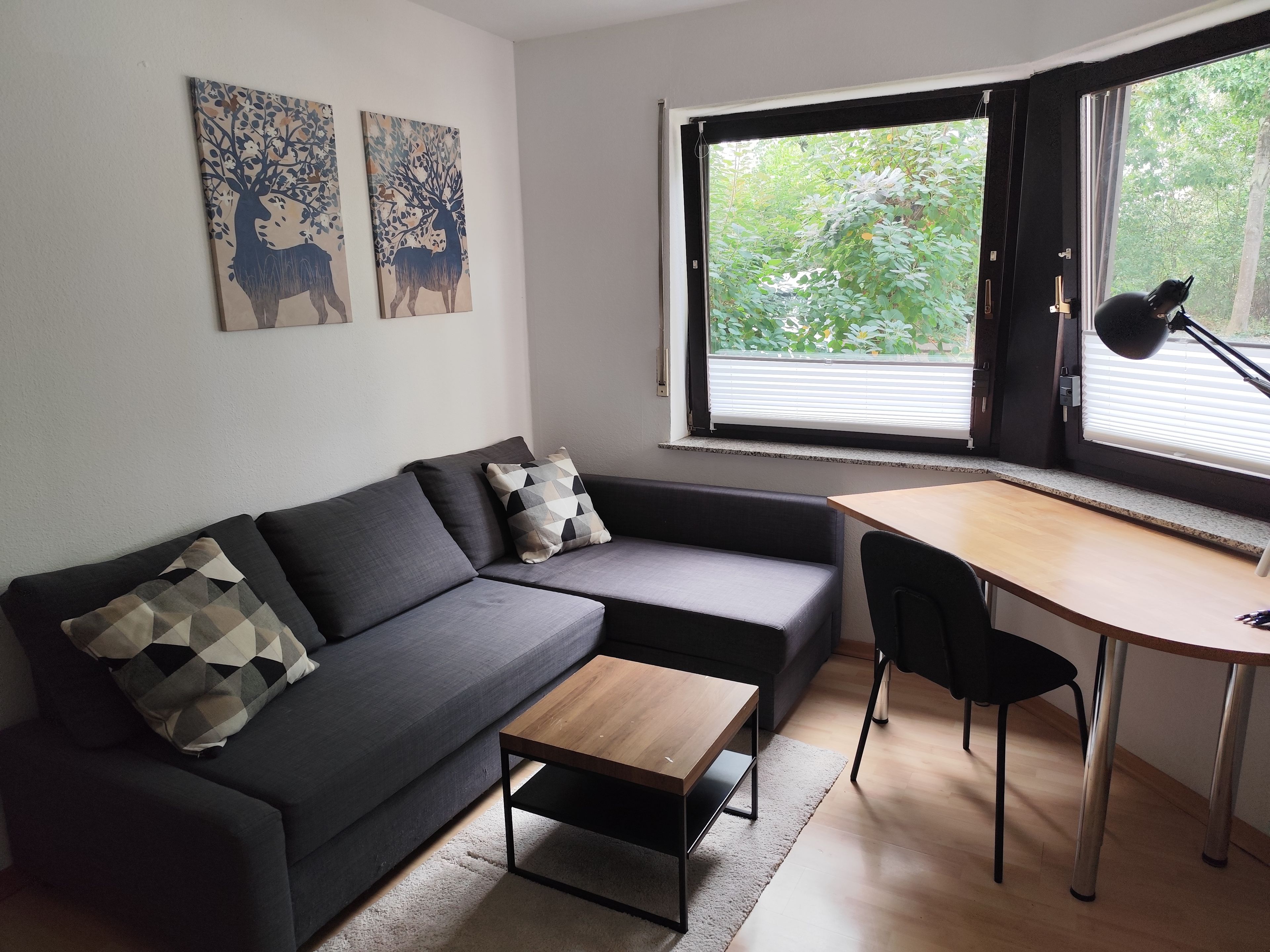 Ein zeitgemäßer Wohnraum mit einem grauen Sofa, einem Holztisch und einem Schreibtisch mit Stuhl. Zwei an der Wand befestigte Kunstwerke zeigen Hirsche und Blattwerk.