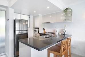 A modern kitchen with a stainless steel refrigerator, a microwave, a stove, and a sink. There are wooden chairs and a plant on the counter. - Couple Escape-10min walk to Hastings St&Beach (Noosa Heads)