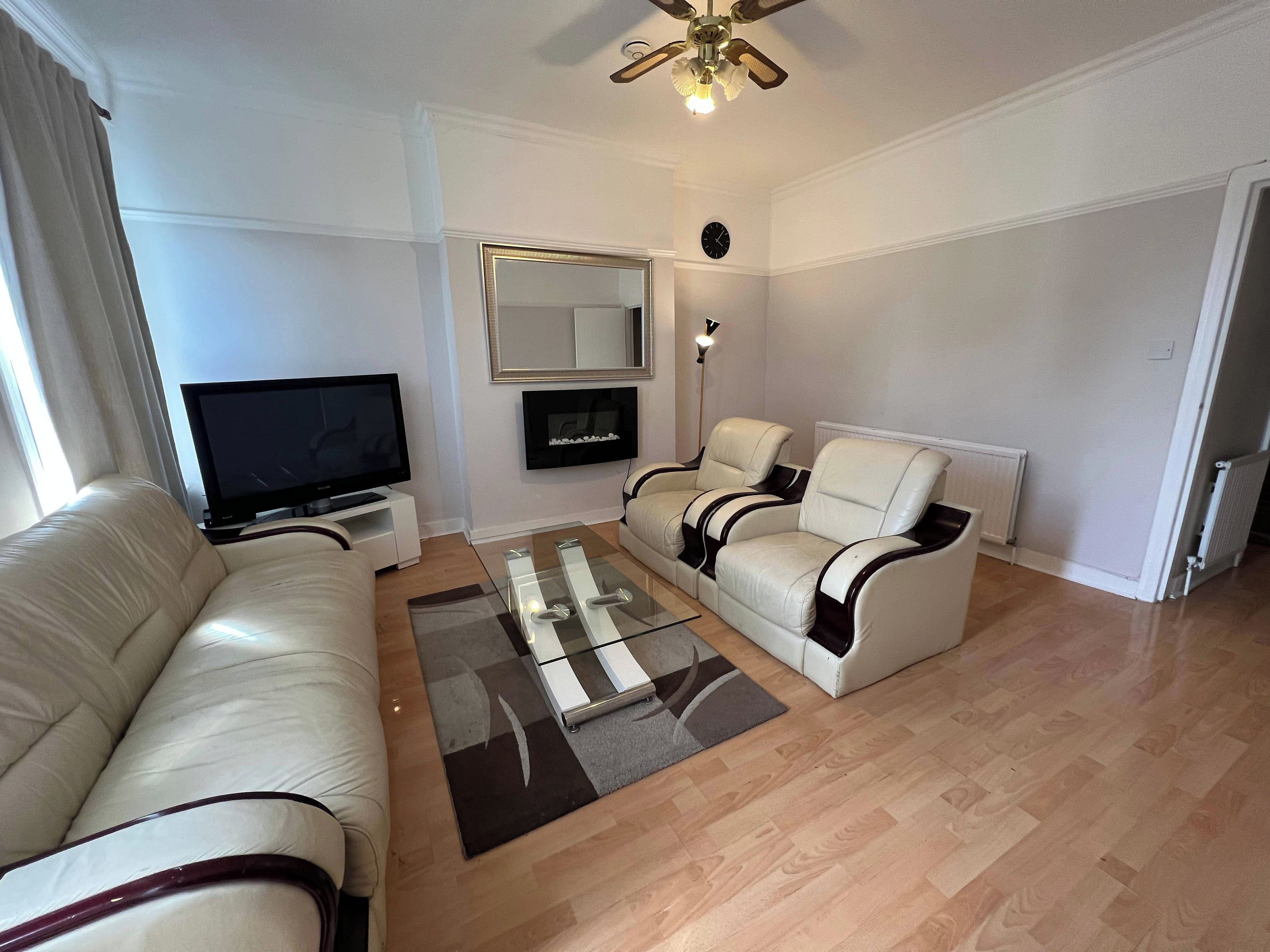 A living room with a sofa, two armchairs, a coffee table, a flat-screen TV, a floor lamp, and a ceiling fan.