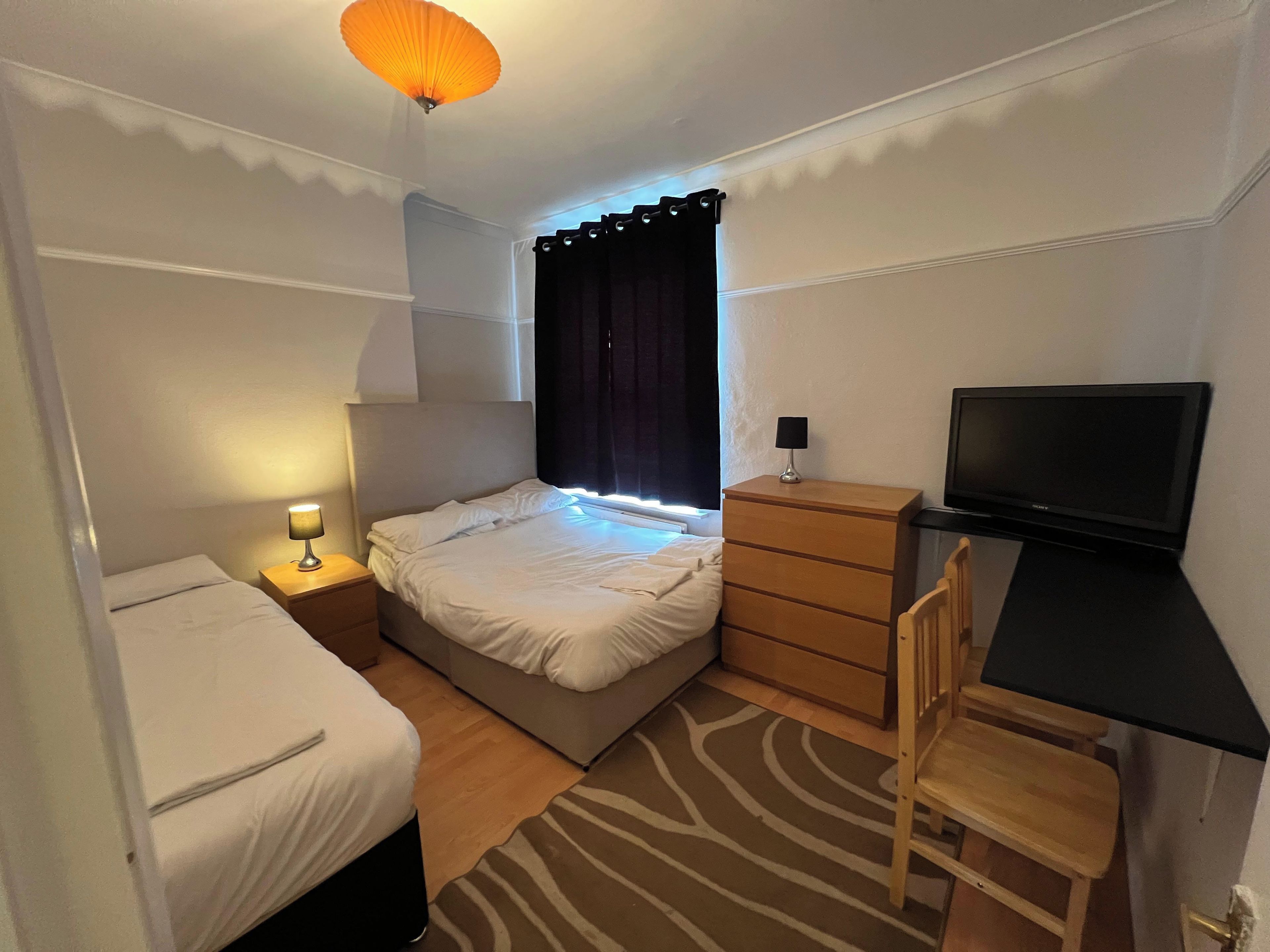 A hotel room with a bed, bedside table with lamp, dresser, desk with chair, and a television.
