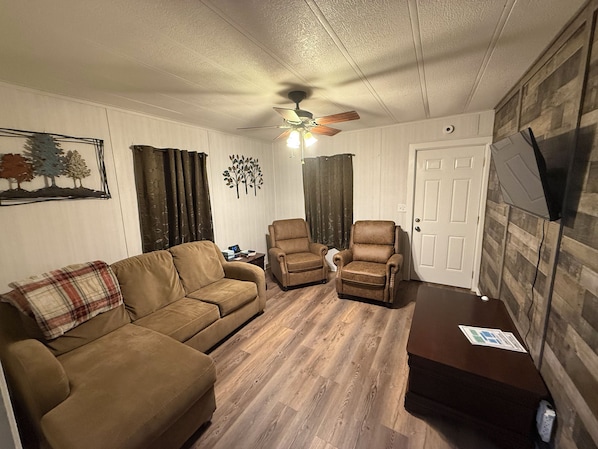 A living room with a brown sofa, two armchairs, a ceiling fan, and a wooden floor.