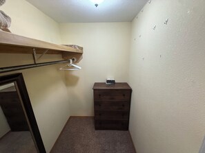 Una habitació petita i estreta amb una prestatgeria de fusta, una barra penjada per a roba, un petit calaix i una porta que dona a una altra habitació.