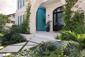 Un bâtiment moderne avec une façade blanche, une entrée en verre bleu et une porte noire. On peut voir une plante en pot sur les marches et diverses plantes ornementales aux alentours.