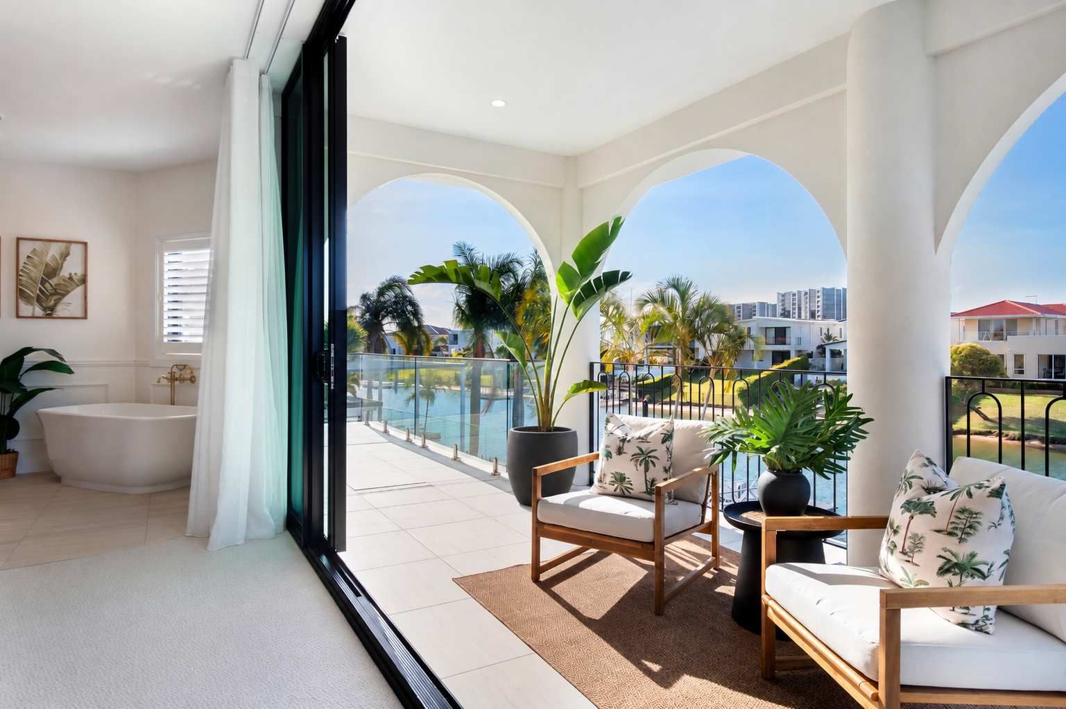 A modern balcony with a view of a swimming pool and palm trees, furnished with a sofa, chairs, and a potted plant.