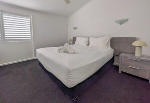 A hotel room with a large bed, a nightstand, a lamp, and a window with blinds. - Ocean Breeze Haven 1 Bed Apartment (Noosa Heads)