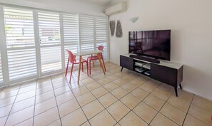 A living room with a TV, a dining table with chairs, and large windows with blinds. - Ocean Breeze Haven 1 Bed Apartment (Noosa Heads)