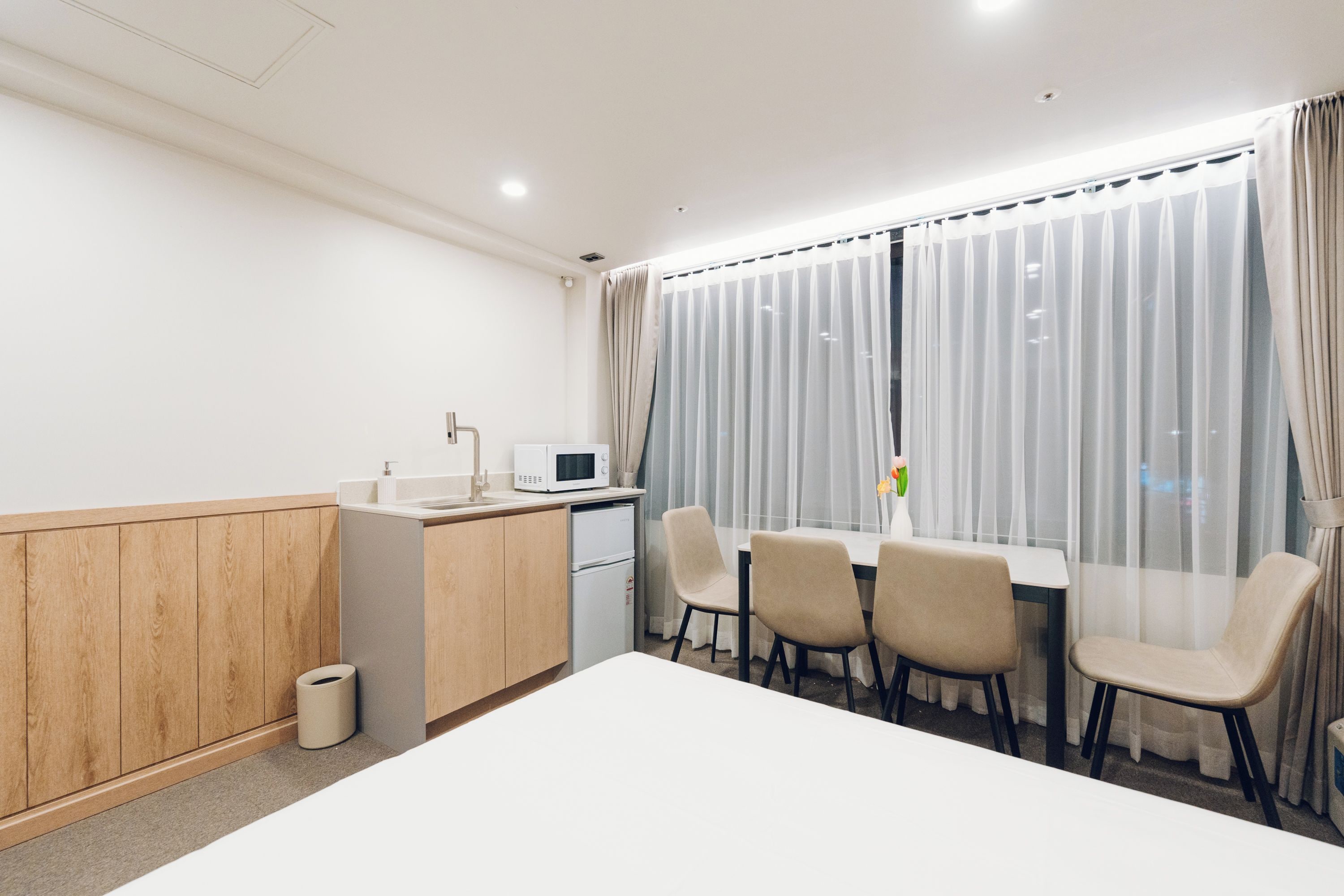 Una habitación de hotel con kitchenette, una mesa de comedor con sillas, un ventanal con cortinas y un cubo de basura.