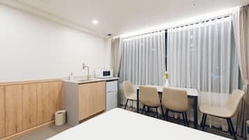 Una habitación de hotel con kitchenette, una mesa de comedor con sillas, un ventanal con cortinas y un cubo de basura.