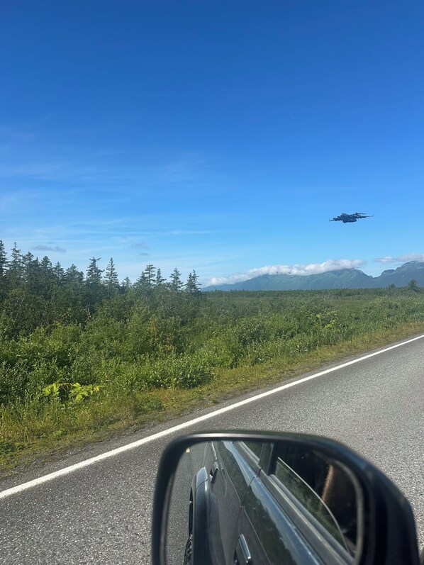 A road with a vehicle's side mirror reflecting a clear sky and a helicopter in flight.