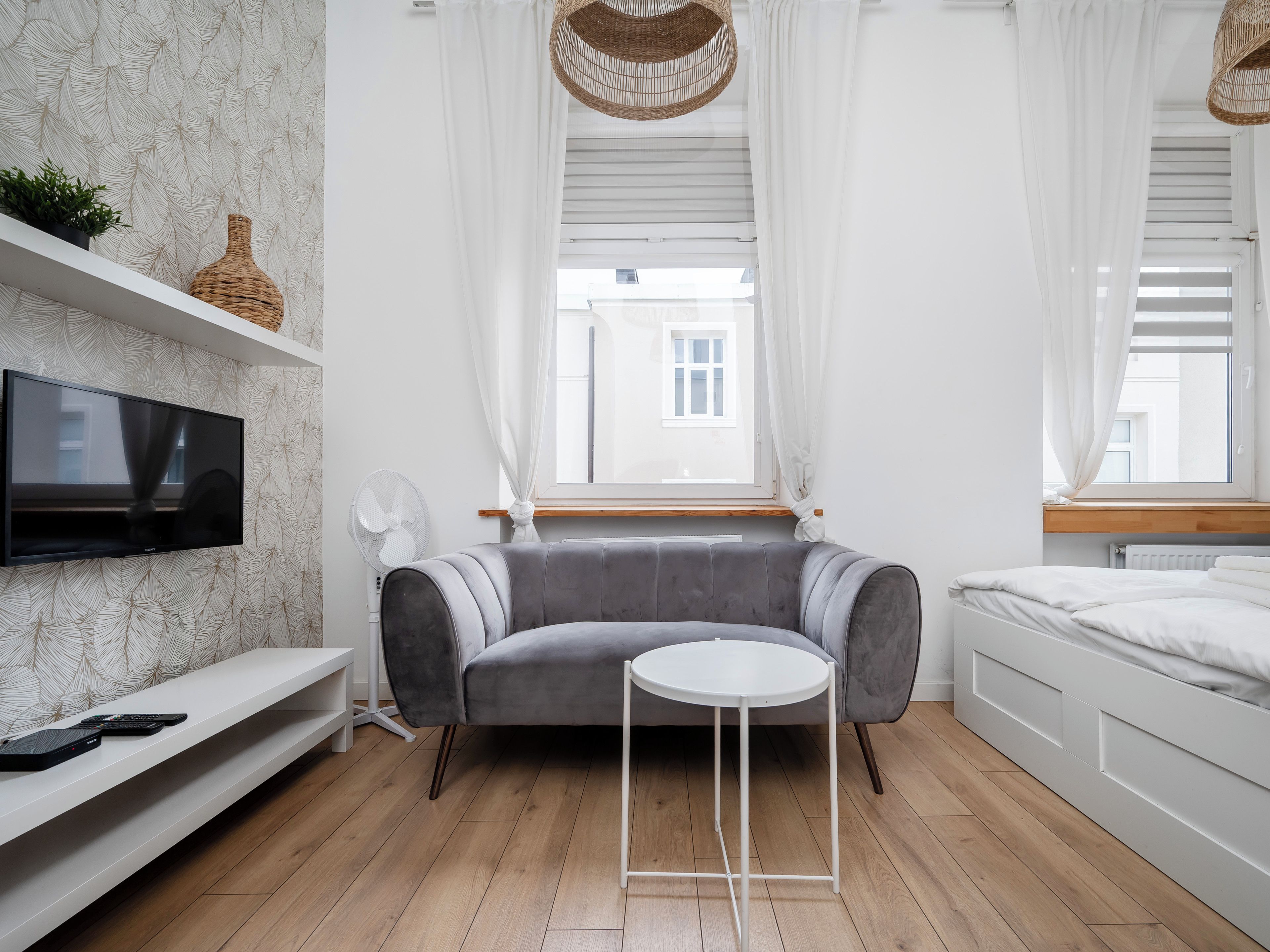 Une chambre moderne avec un canapé gris, une petite table ronde, un téléviseur à écran plat et une fenêtre avec des rideaux blancs.