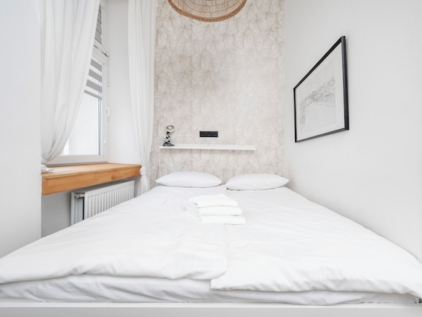 Une chambre à coucher au style minimaliste, avec un lit, du linge de lit blanc, une table de chevet, une étagère fixée au mur, un tableau encadré et une fenêtre avec des rideaux.