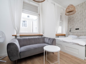 A modern bedroom with a grey sofa, a small round table, a bed with white bedding, and a window with white curtains. - Narutowicza 41 Boutique Apartment #2 (Łódź)