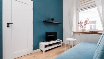 A modern living room with a teal accent wall, a white door, a flat-screen TV on a white stand, a blue sofa, and a small white side table.