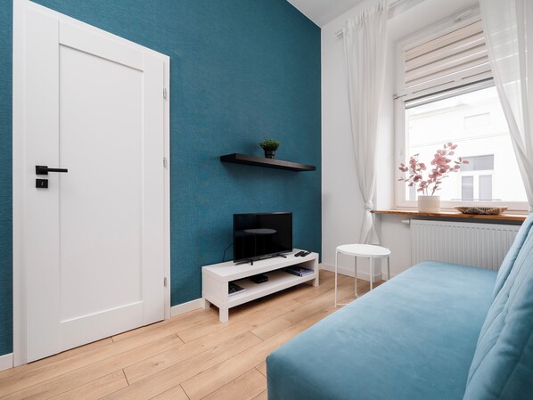 Een moderne woonkamer met een turquoise accentmuur, een witte deur, een flatscreen-televisie op een wit meubelstuk, een blauwe bank en een klein wit bijzettafeltje.