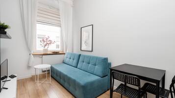 A modern living room with a teal sofa, a small round table, a black dining table with chairs, a wicker ceiling light, and a framed picture on the wall.