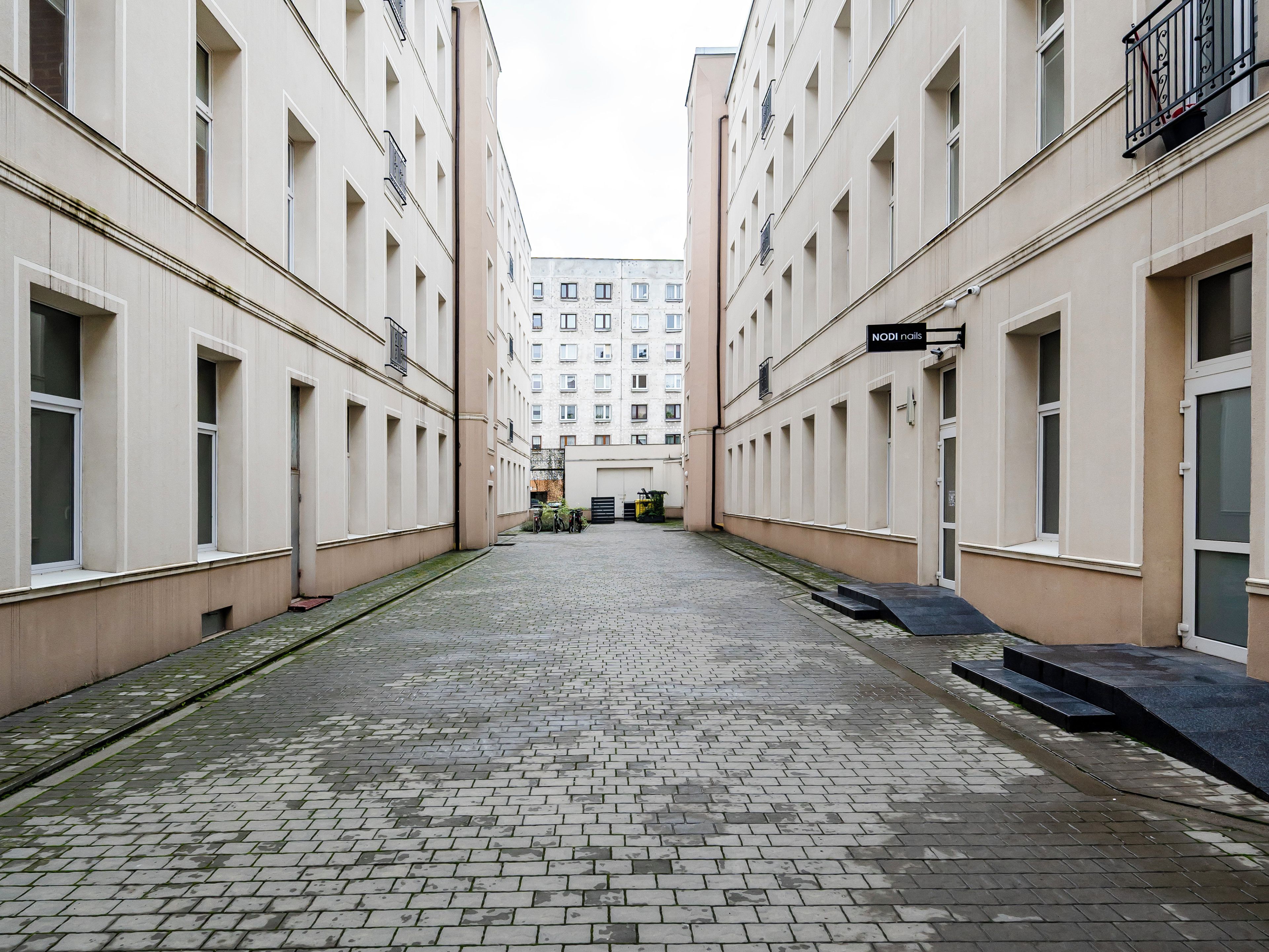 A narrow alleyway between buildings with a paved ground, a sign for 'NODI nails', and a bicycle parked on the right.